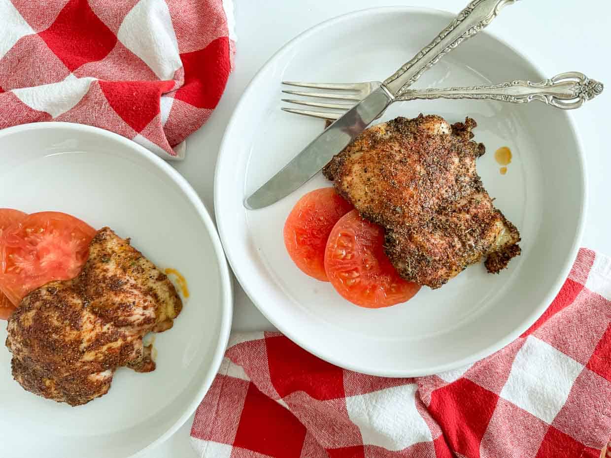 Two plates with seasoned, cooked chicken thighs and sliced tomatoes, placed on a white surface with red and white checkered napkins. Fork and knife rest on one plate.