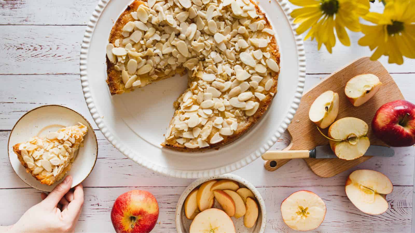 A sliced almond apple cake on a white plate, with a piece served on a small dish. Surrounding are apples, apple slices, a knife, and yellow flowers on a wooden table.