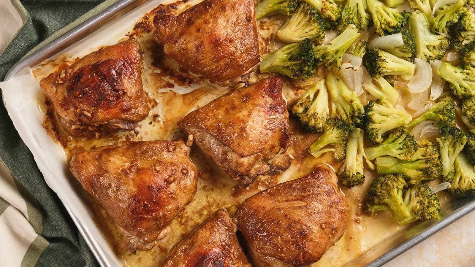 Baked chicken thighs and roasted broccoli with onions arranged on a parchment-lined baking sheet.