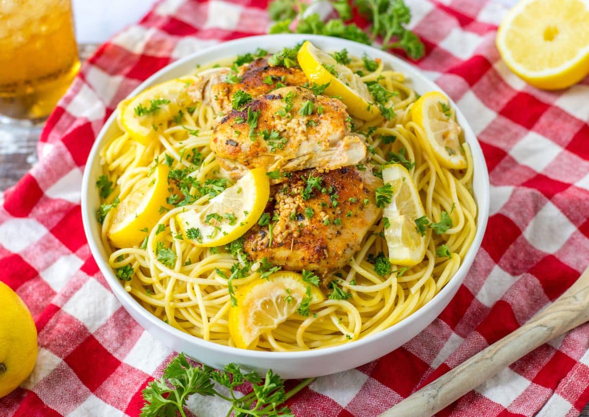 A bowl of spaghetti topped with roasted chicken, lemon slices, and chopped parsley sits on a red and white checkered tablecloth.