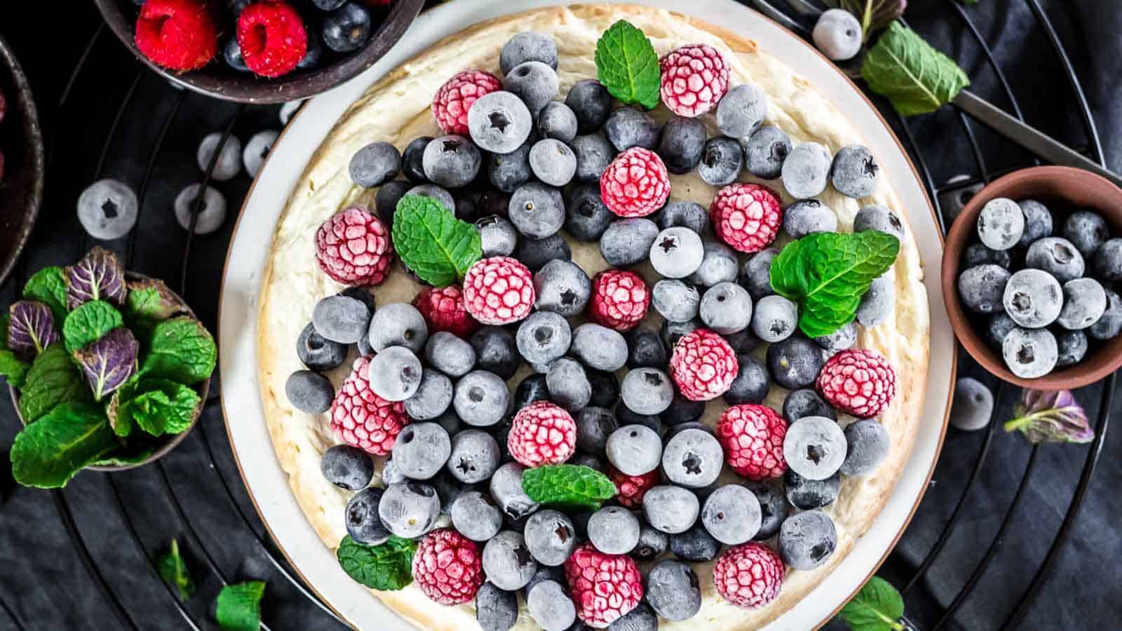 A cheesecake topped with fresh blueberries, raspberries, and mint leaves, surrounded by bowls of berries and mint on a black surface.