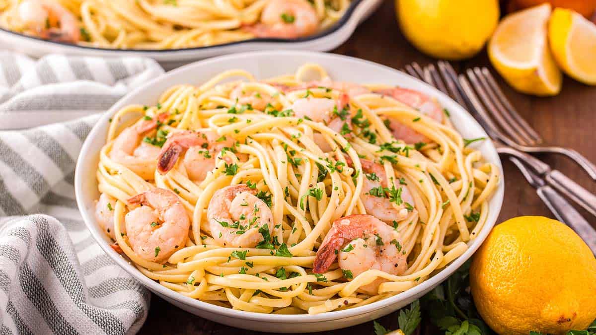 A bowl of shrimp pasta garnished with chopped parsley, with lemons, forks, and a striped napkin on a wooden table.