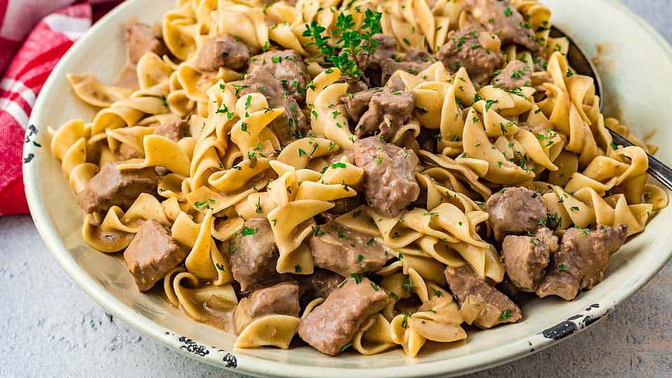 A plate of beef stroganoff with egg noodles, chunks of beef, creamy sauce, and garnished with chopped parsley.
