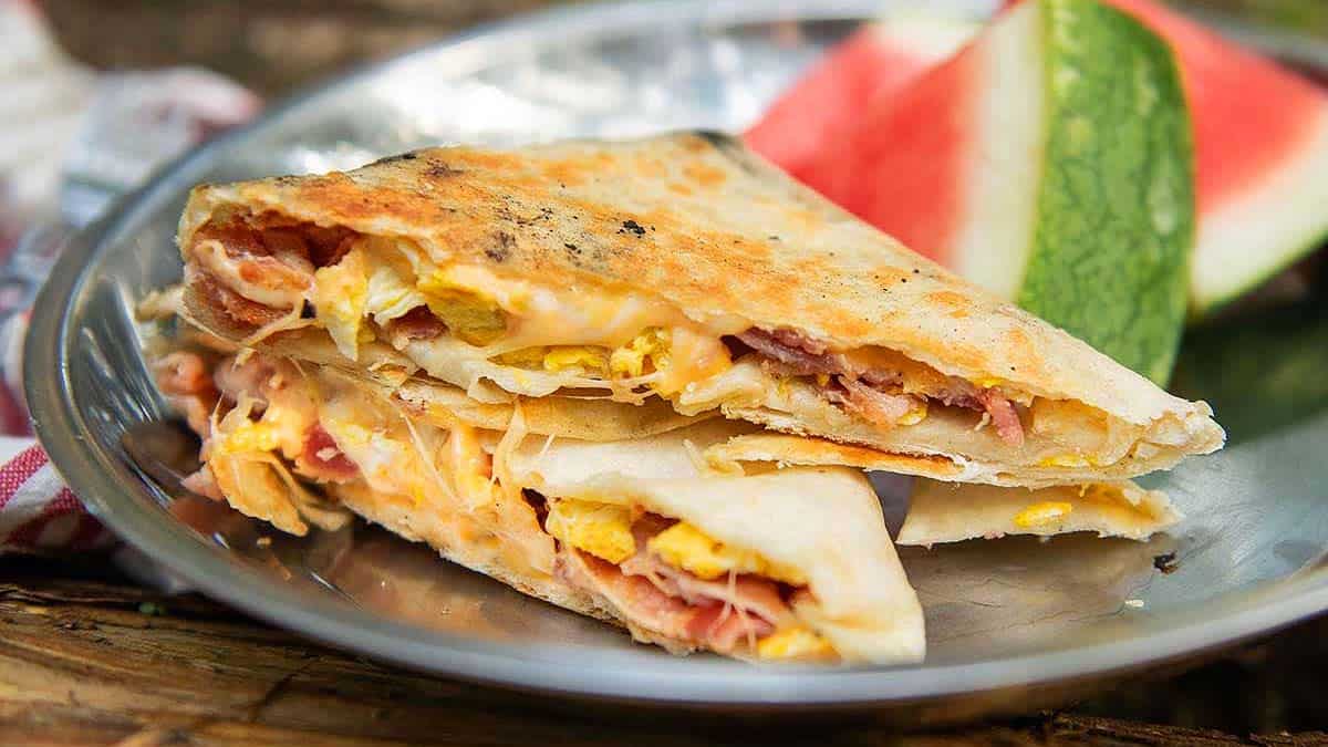 Two quesadilla wedges filled with eggs, cheese, and bacon on a plate, served with a slice of watermelon in the background.