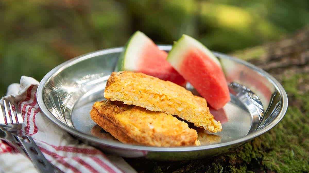 A metal plate holds two slices of cornbread and two wedges of watermelon, with a striped napkin and forks beside it.