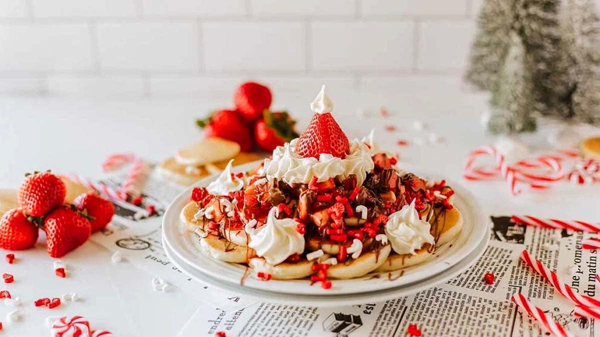 A plate of waffles topped with whipped cream, strawberries, chocolate drizzle, and crushed candy canes sits on a table with strawberries and holiday decorations in the background.