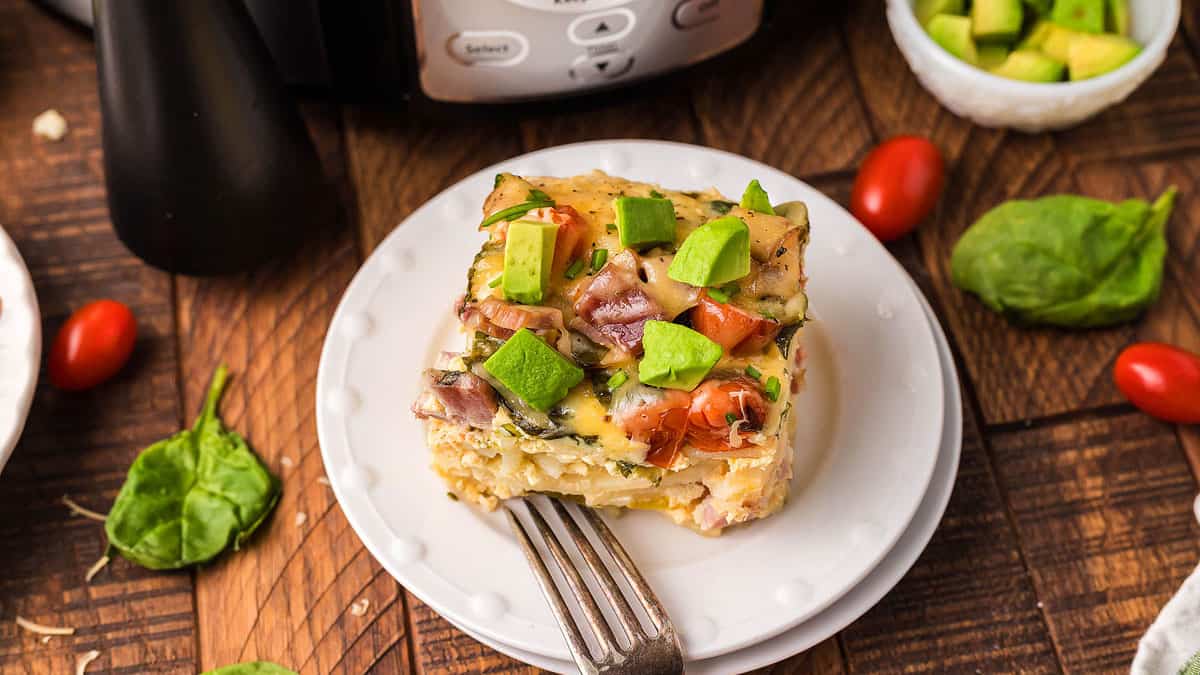 A slice of casserole topped with diced avocado sits on a white plate with a fork; spinach leaves and cherry tomatoes are scattered nearby.