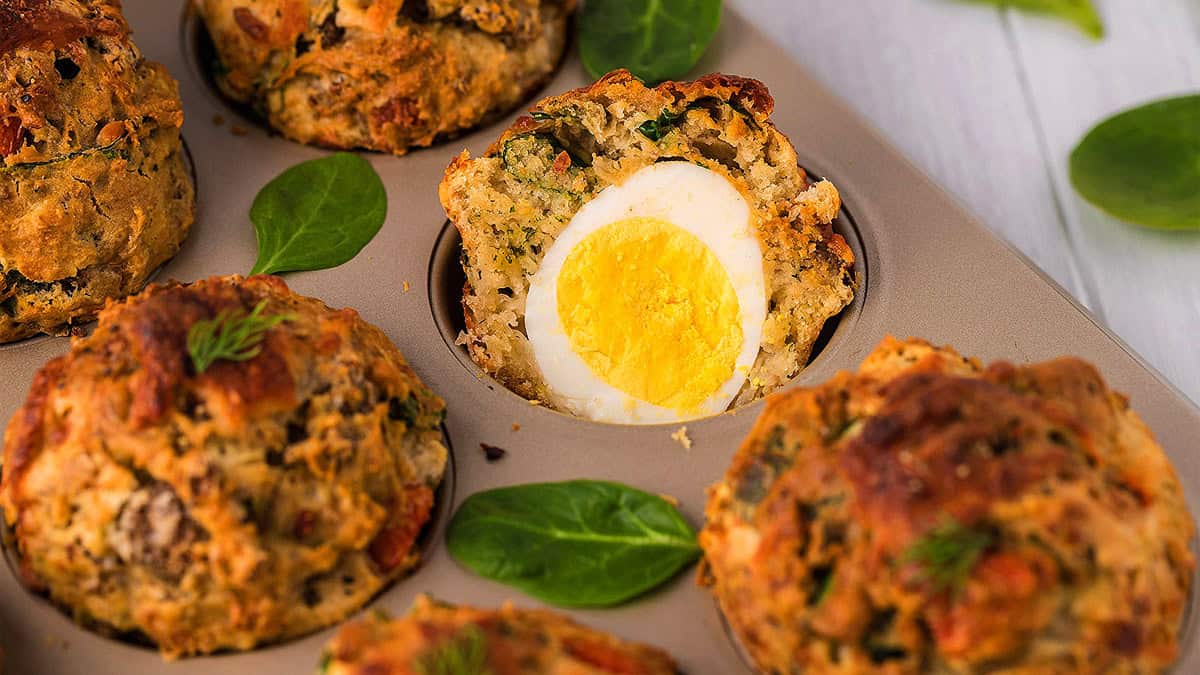 Muffin tin with savory baked muffins, one cut open to reveal a whole hard-boiled egg inside; spinach leaves scattered around.