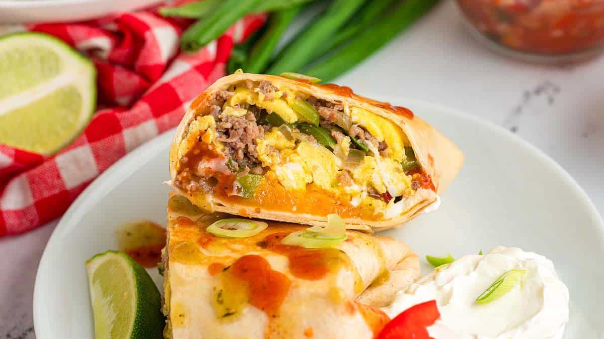 A breakfast burrito cut in half showing eggs, beef, cheese, and vegetables, served with sour cream, green onions, and lime on a white plate.
