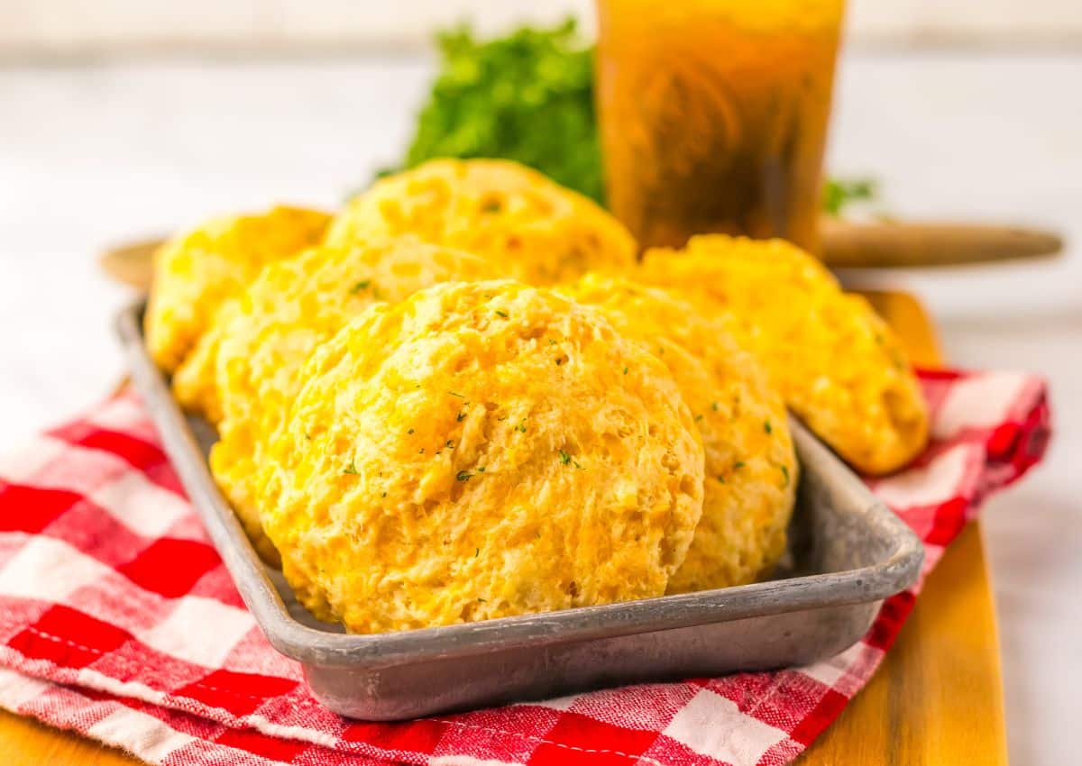 A metal tray holds several golden cheddar biscuits, placed on a red and white checkered cloth, with a drink and green herbs in the background.