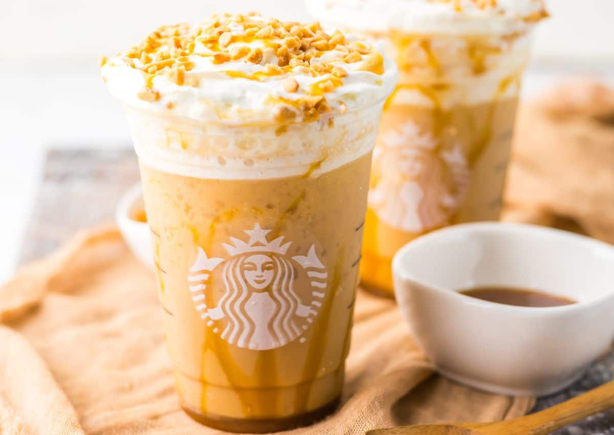 A Starbucks cold blended drink topped with whipped cream and chopped nuts, served in a clear cup with caramel drizzle, next to a small white bowl of sauce.