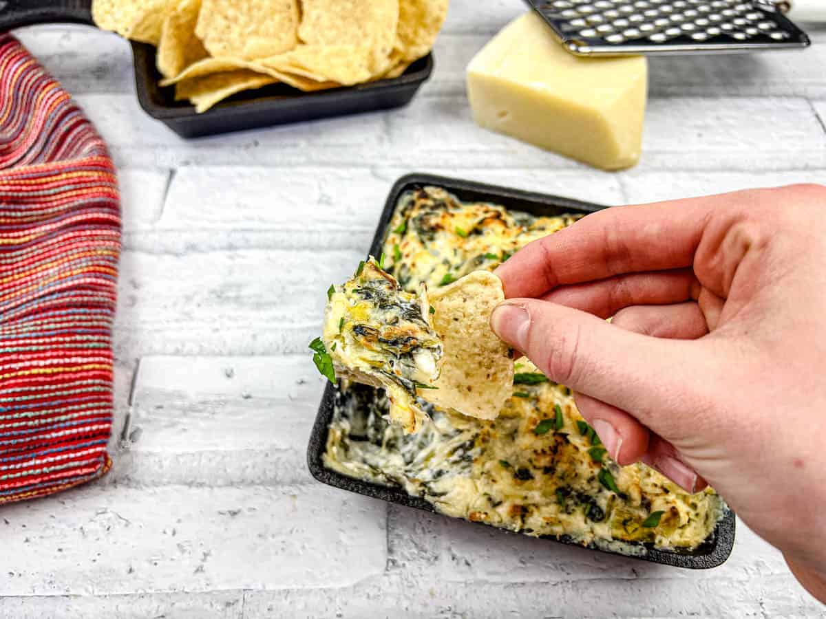 A hand dips a tortilla chip into a dish of baked spinach artichoke dip, with a wedge of cheese and more chips in the background.