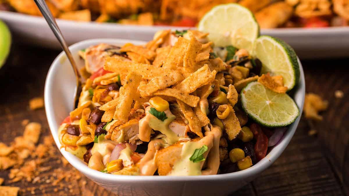 A bowl of taco salad topped with corn, crushed tortilla chips, creamy dressing, and garnished with lime slices.