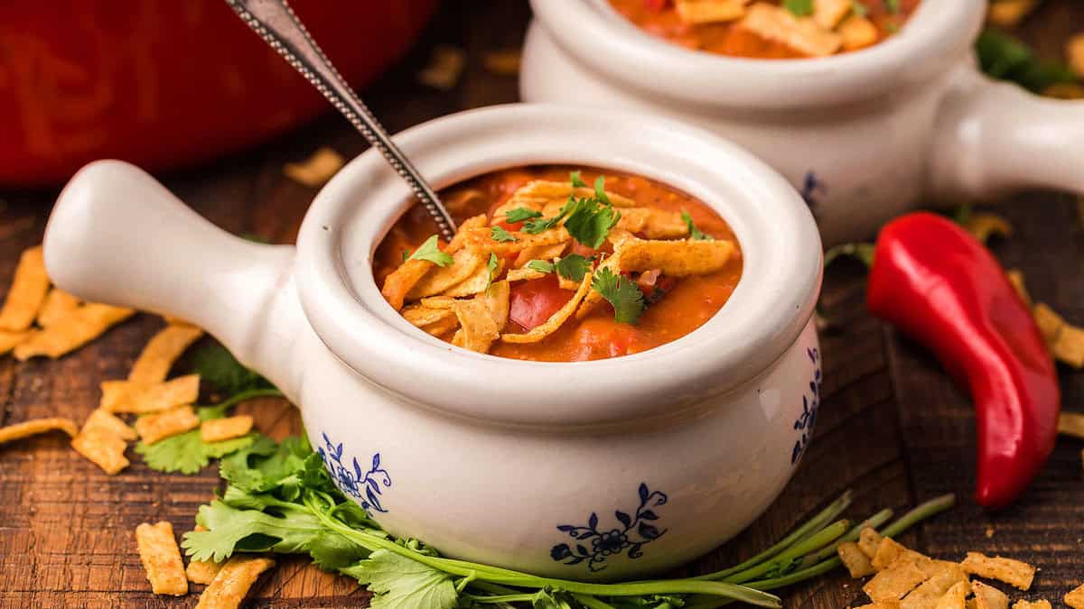 A bowl of soup garnished with tortilla strips and cilantro, with a spoon inside. Fresh cilantro and a red chili pepper are on the wooden table nearby.