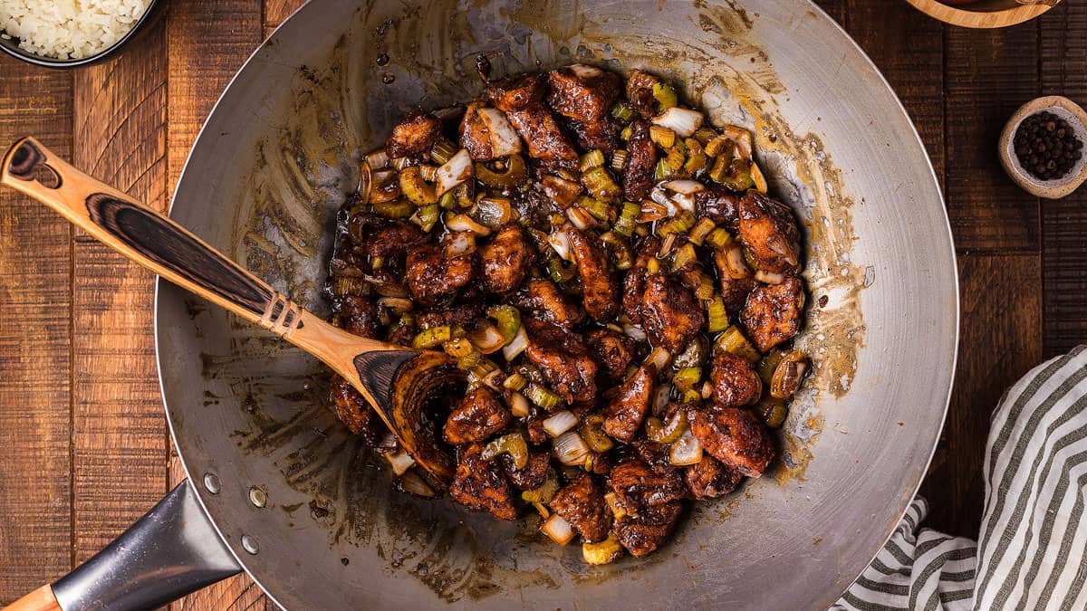 A wok filled with stir-fried chicken, diced onions, and green peppers being mixed with a wooden spoon on a wooden table.