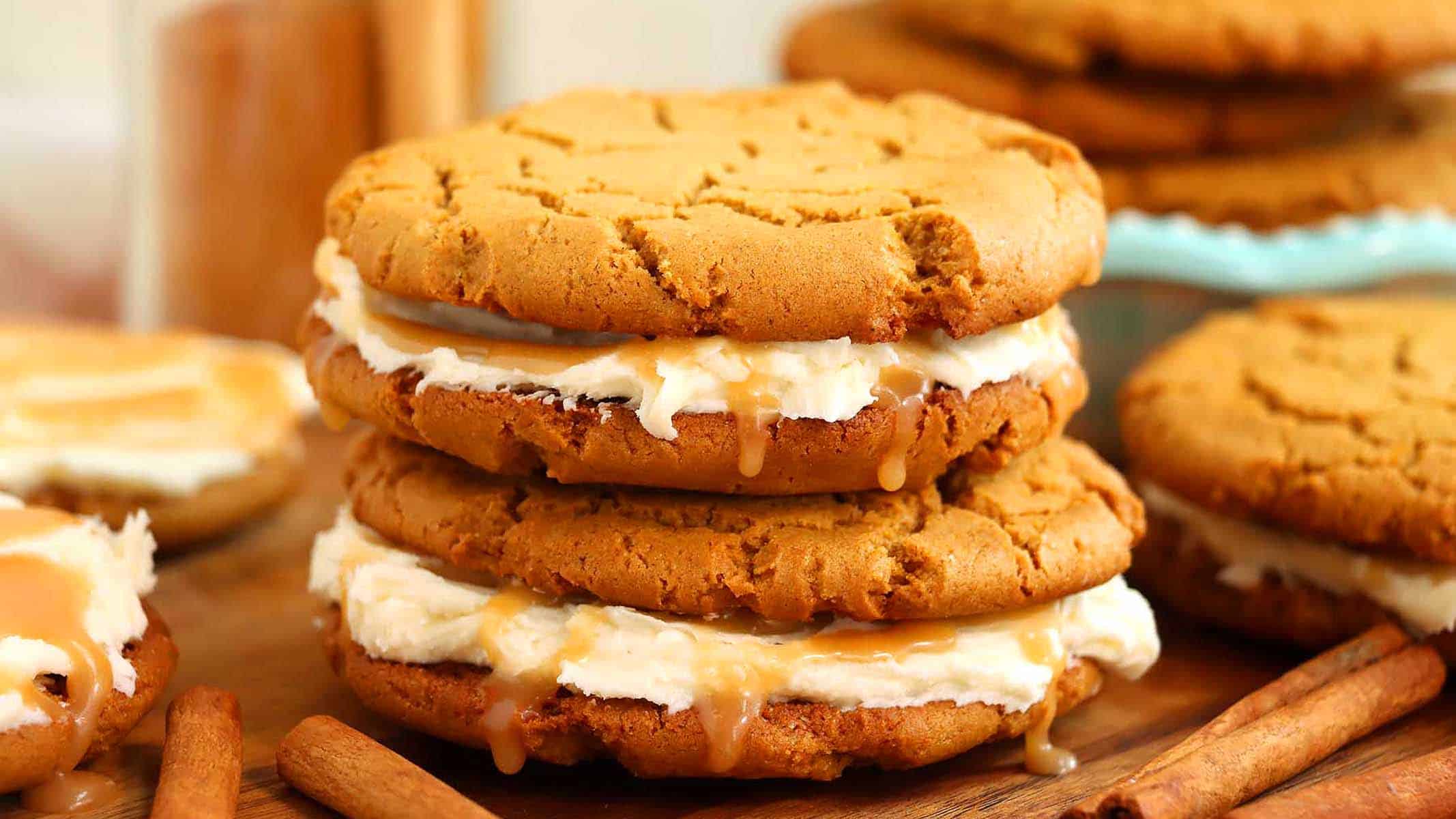 Two caramel-drizzled cookie sandwiches filled with cream frosting are stacked on a wooden surface, with cinnamon sticks and more cookies in the background.