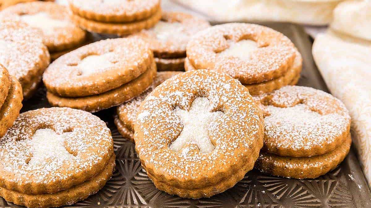 Round sandwich cookies with powdered sugar dusting, each featuring a cut-out in the shape of a gingerbread person, arranged on a patterned tray.