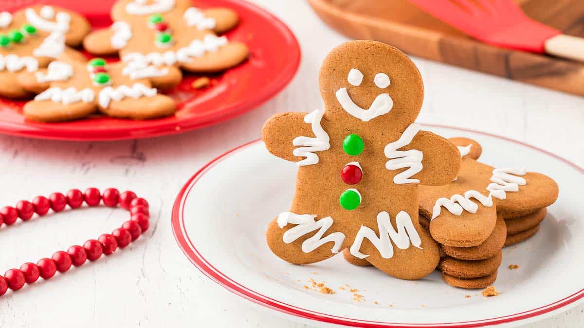 A plate of gingerbread cookies shaped like people, decorated with white icing and red and green candies, sits on a table next to another plate of cookies.
