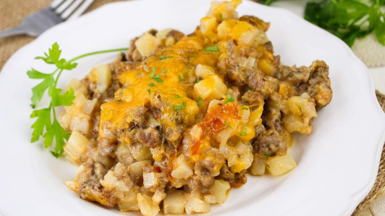 A serving of cheesy hashbrown casserole with ground beef on a white plate, garnished with a sprig of parsley.