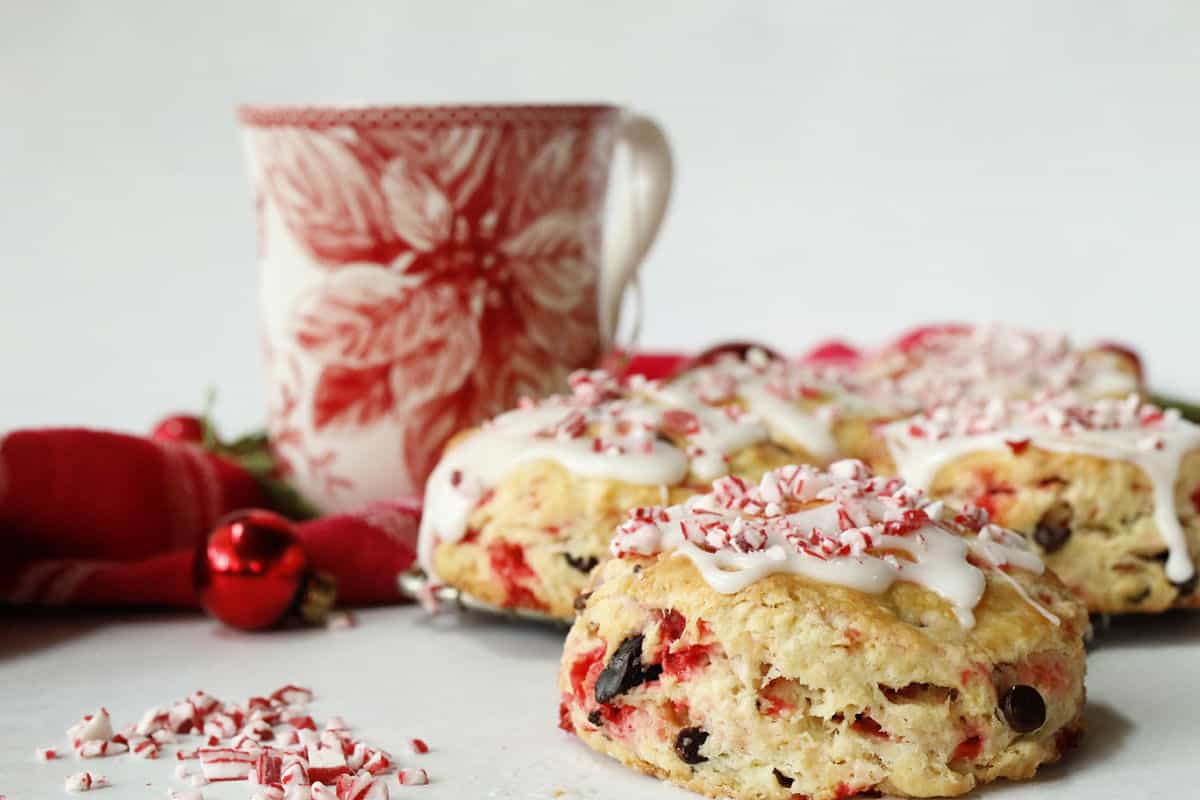 A close-up of peppermint scones topped with icing and crushed candy, with a festive mug and holiday decorations in the background.
