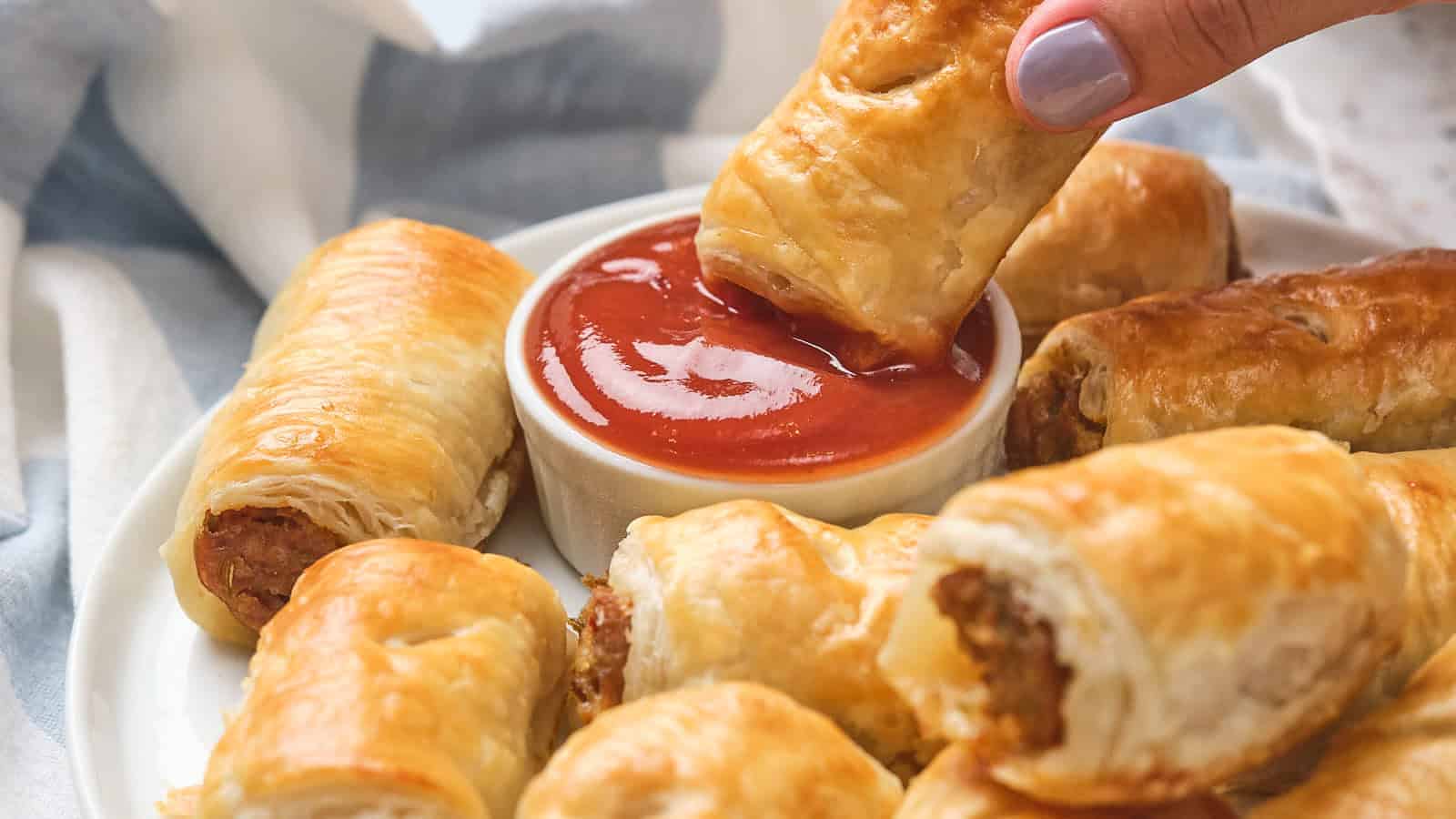 A hand dips a sausage roll into a bowl of tomato sauce, surrounded by more sausage rolls on a white plate.