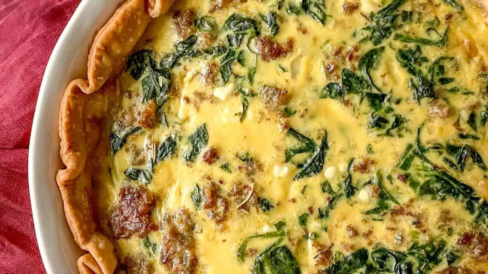 Close-up of a baked quiche with a golden crust, visible pieces of sausage, spinach, and egg mixture in a white dish.