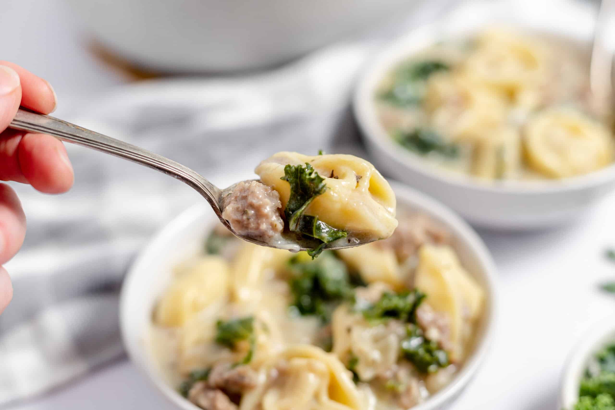 A close-up of a spoon holding tortellini, sausage, and kale over a bowl of creamy soup.