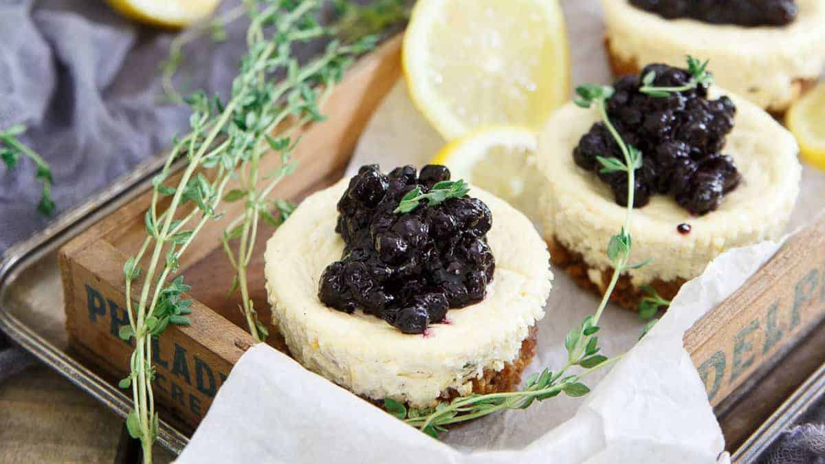 Mini cheesecakes topped with blueberry compote, garnished with fresh thyme, and served with lemon slices on a tray.