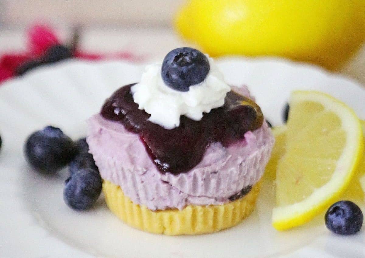 A mini blueberry cheesecake topped with blueberry sauce, whipped cream, and a blueberry, served with fresh blueberries and a lemon wedge on a white plate.
