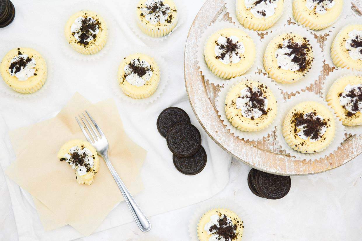 Mini cheesecakes topped with crushed chocolate cookies are arranged on a cake stand and a napkin, with whole cookies and a fork nearby.