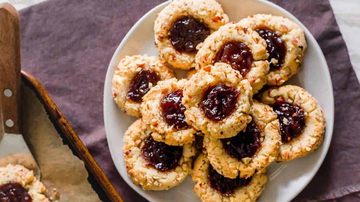 A plate of thumbprint cookies filled with red jam, surrounded by a few more cookies on a baking tray lined with parchment paper.