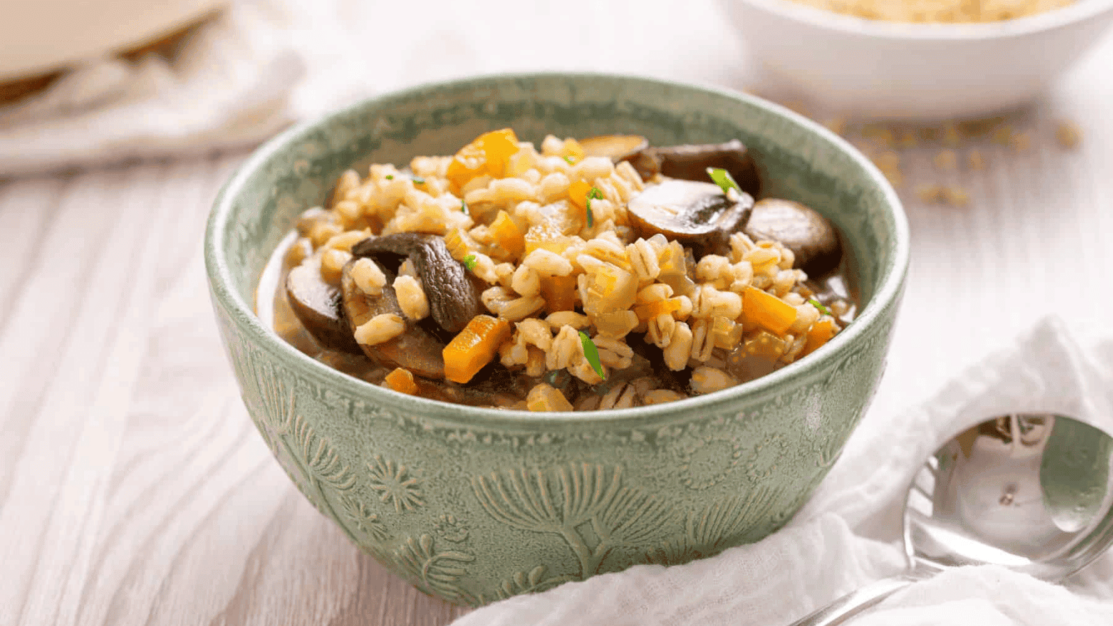 A green textured bowl filled with barley soup containing mushrooms, carrots, and herbs, placed on a light-colored table next to a spoon and napkin.