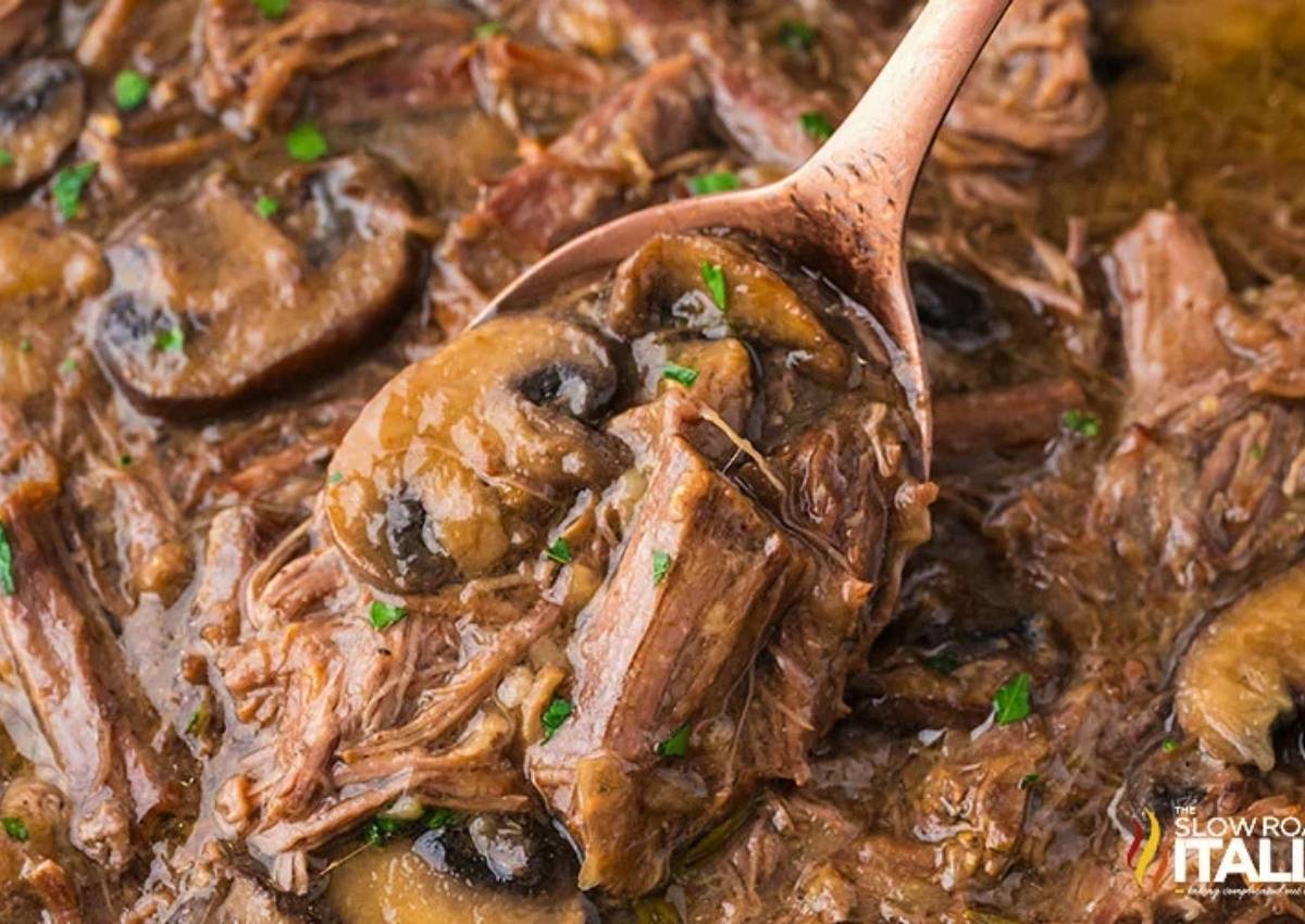 A close-up of a spoonful of beef pot roast with mushrooms in a thick brown gravy, garnished with chopped herbs.