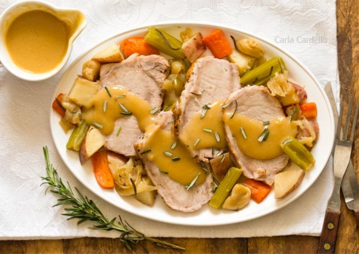 A platter of sliced roast pork topped with gravy, served over roasted vegetables including carrots, potatoes, and celery, with a small bowl of gravy and a sprig of rosemary nearby.