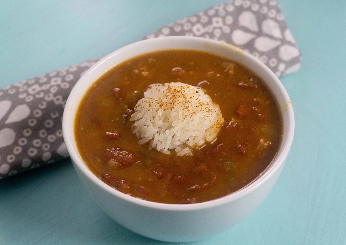 A bowl of gumbo with a mound of white rice in the center, placed on a light blue surface next to a folded gray and white patterned napkin.