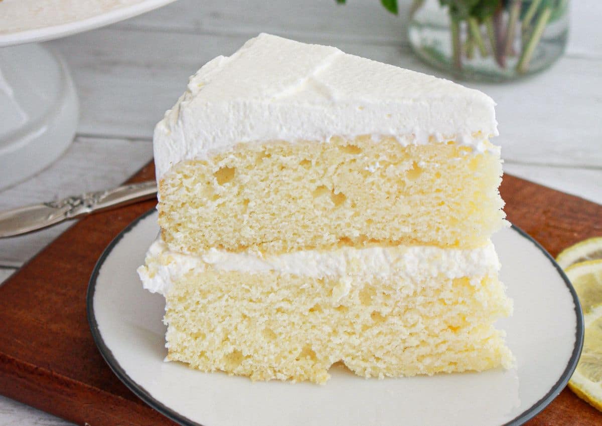 A slice of two-layer vanilla cake with white frosting on a white plate, placed on a wooden board.