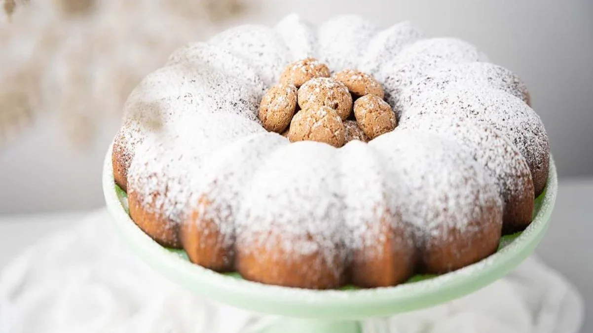 A bundt cake topped with powdered sugar and small round cookies is displayed on a light green cake stand.