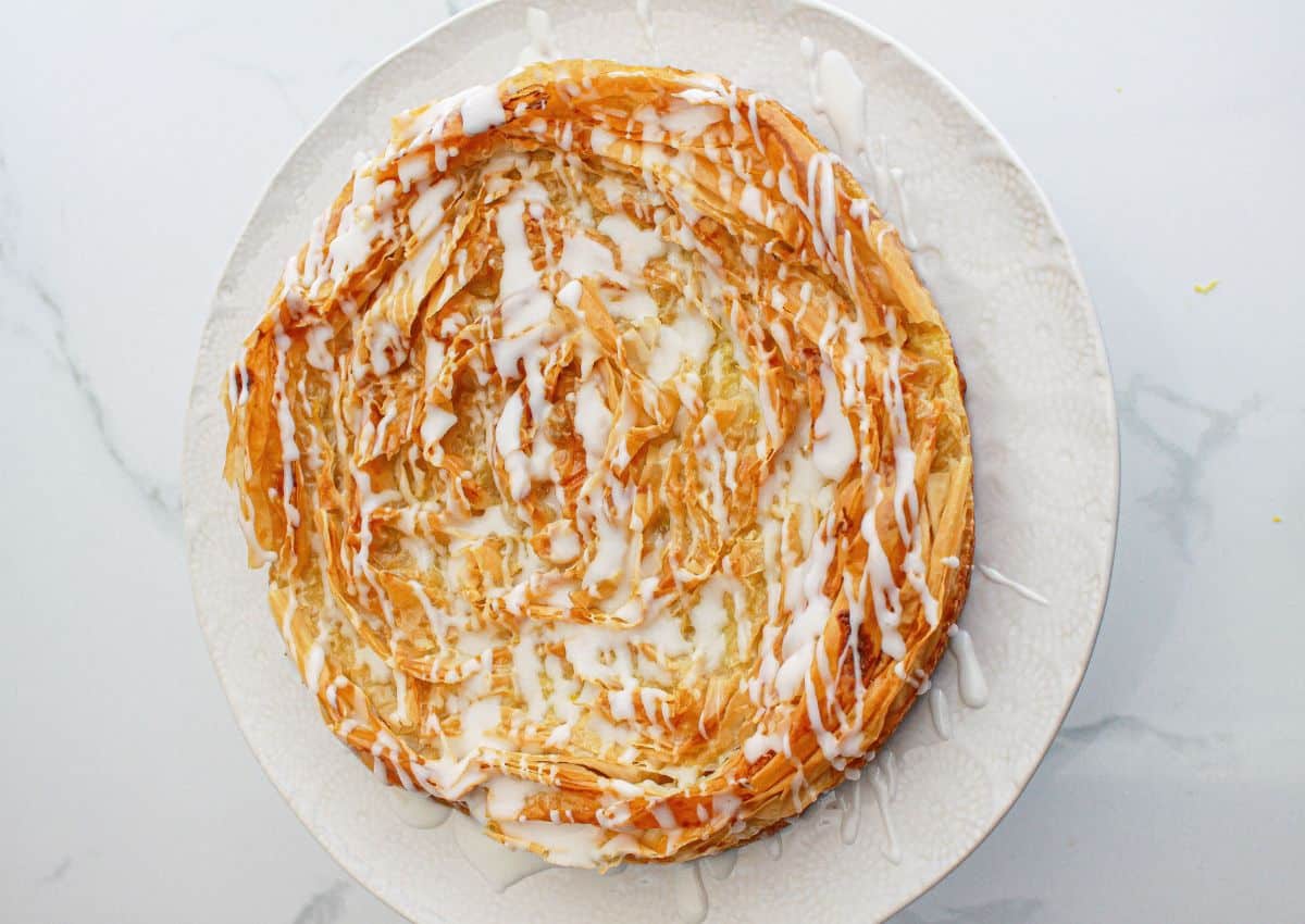 A round pastry with layered, golden flaky crust is topped with a white icing drizzle, served on a white plate.
