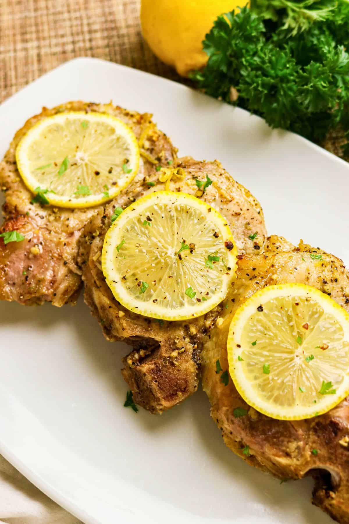 Three cooked pork chops garnished with lemon slices and herbs are arranged on a white plate with parsley and a lemon in the background.
