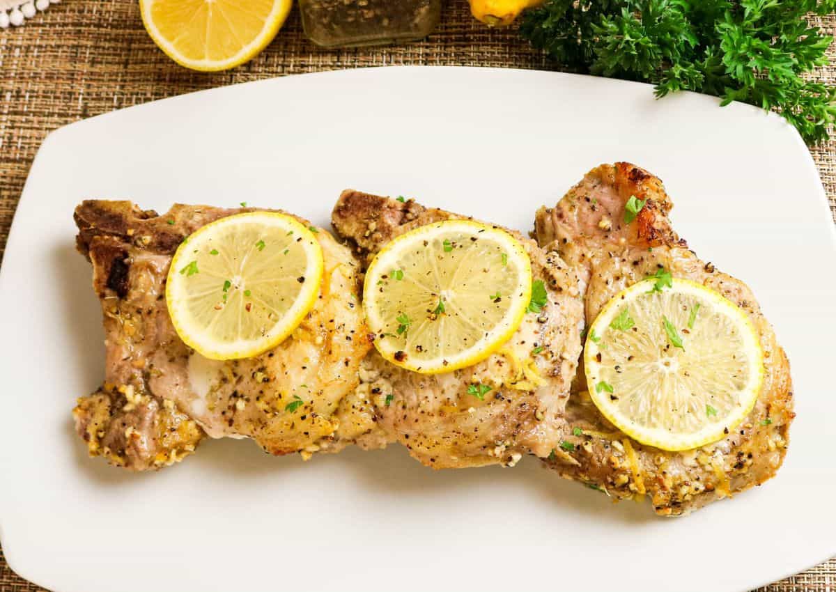 Three cooked pork chops garnished with lemon slices and herbs, served on a white rectangular plate.