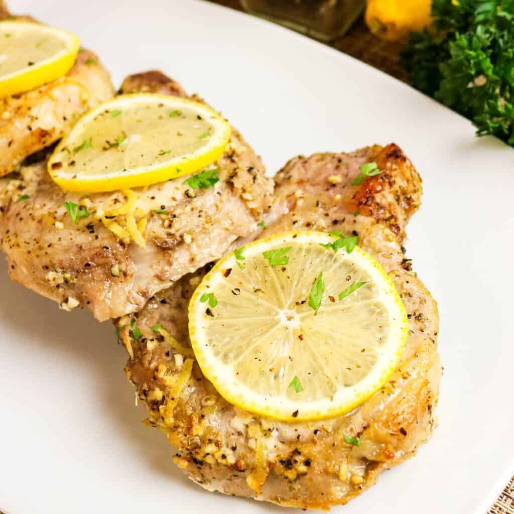 Two cooked pork chops seasoned with herbs and topped with lemon slices are served on a white plate.