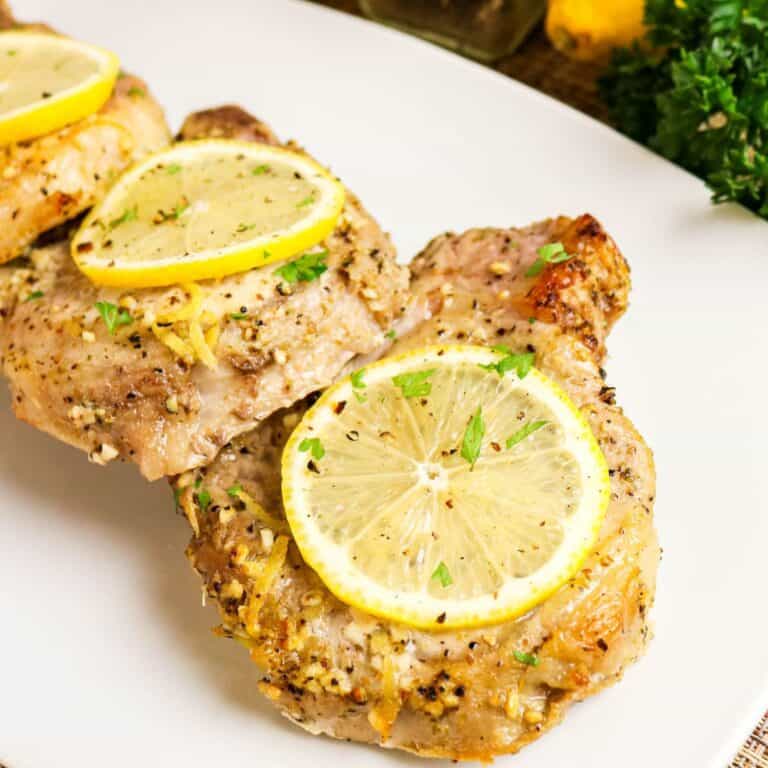 Two cooked pork chops seasoned with herbs and topped with lemon slices are served on a white plate.