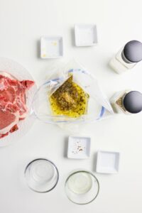 Overhead view of raw pork chops on a plate, a plastic bag with marinade, empty spice jars, and small bowls on a white surface.
