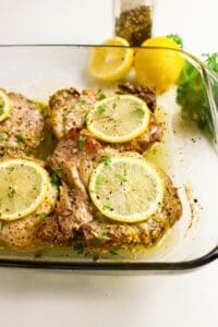 Baked pork chops in a glass dish, topped with lemon slices and herbs, with whole lemons and a pepper grinder in the background.