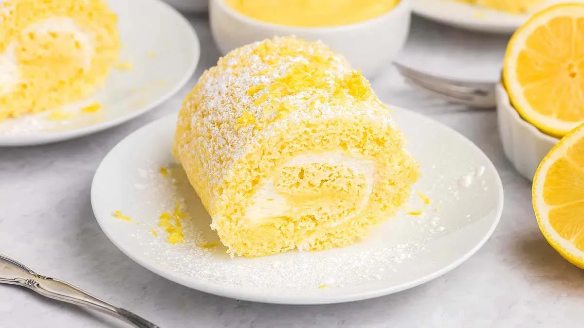 A slice of lemon Swiss roll cake dusted with powdered sugar on a white plate, with lemon halves and bowls in the background.