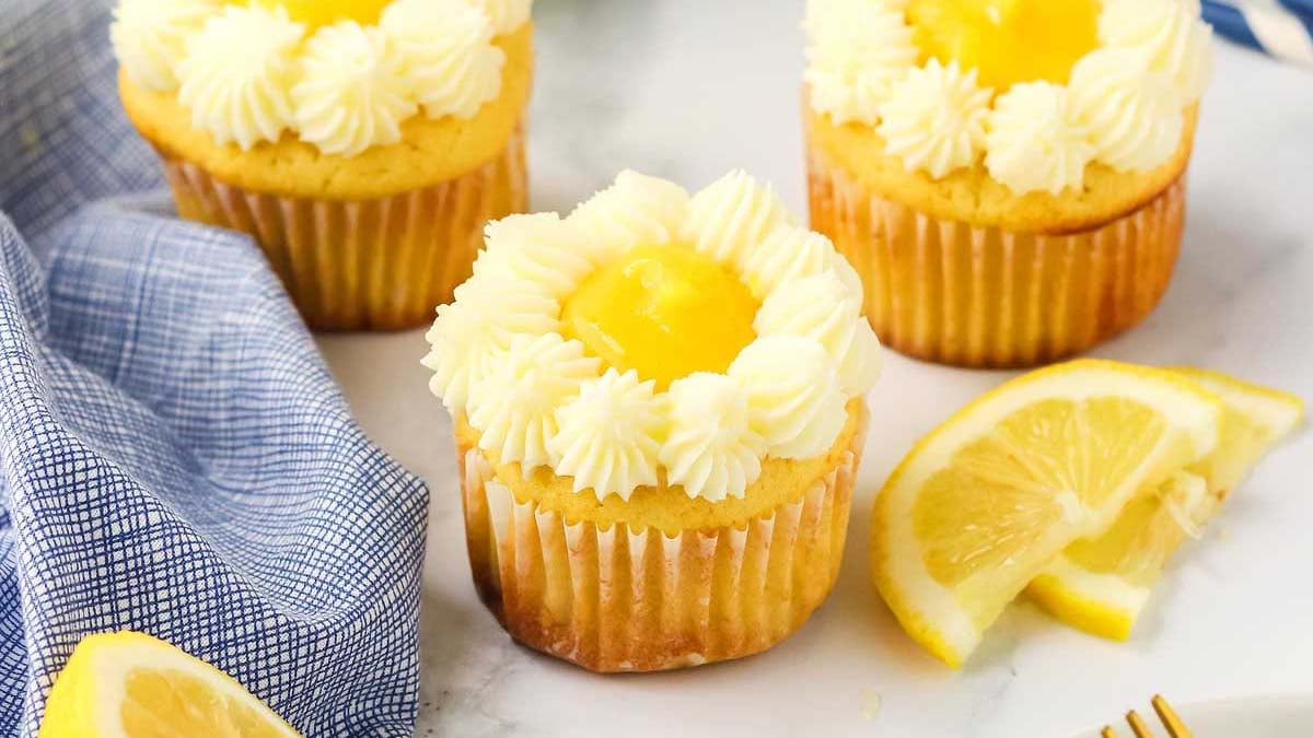 Three lemon cupcakes with white frosting and lemon curd centers are arranged on a white surface, with lemon slices and a blue checked cloth nearby.