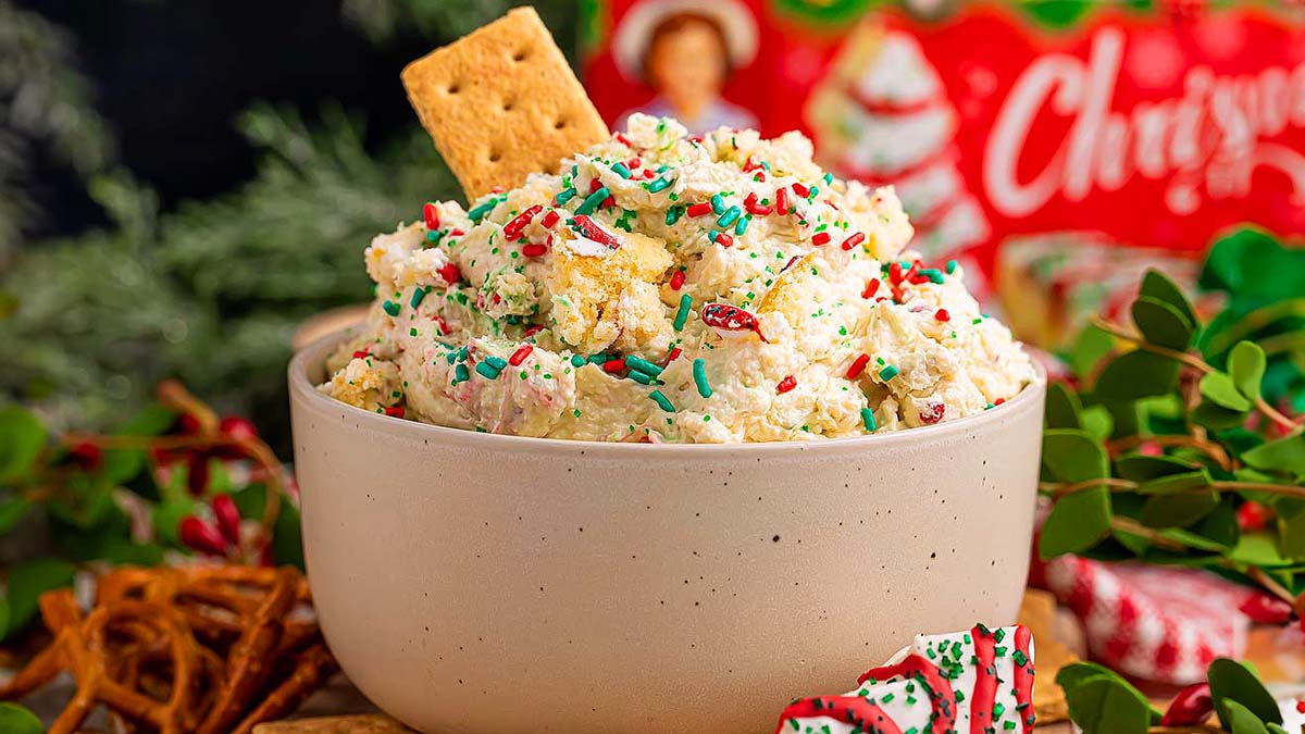 A bowl of festive dessert dip topped with colorful sprinkles and a graham cracker, surrounded by pretzels and holiday decorations.