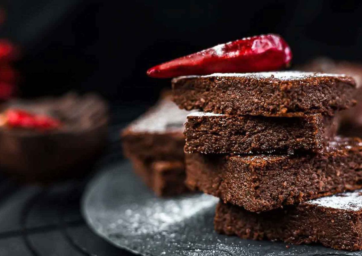 A stack of chocolate brownies on a dark plate, topped with a red chili pepper, with powdered sugar sprinkled on the surface.