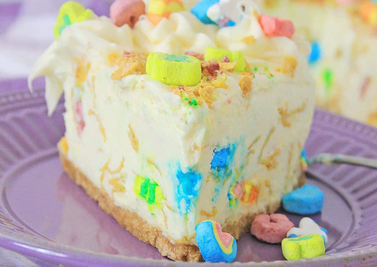 A slice of cheesecake with a crumb crust, colorful cereal pieces mixed in and on top, served on a purple plate.