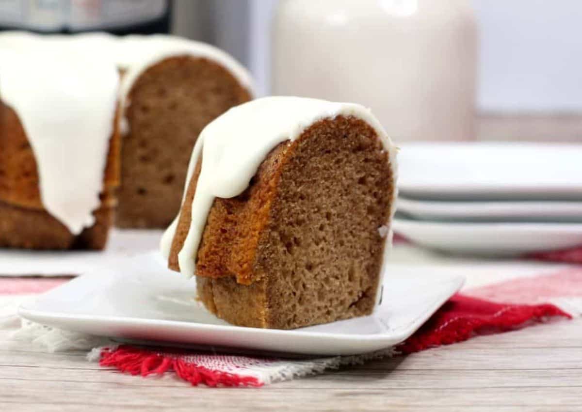 A slice of bundt cake with white icing sits on a white plate, with more cake and stacked plates in the background.